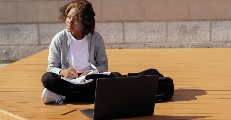 Online Platform - Contemplative African American schoolkid with copybook and netbook sitting with crossed legs on wooden platform while looking away in sunlight