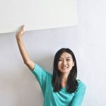 Drones Real Estate - Cheerful Asian woman sitting cross legged on floor against white wall in empty apartment and showing white blank banner